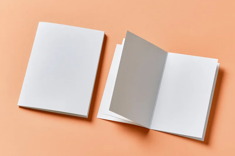Two blank white bifold brochures, one open and one closed, on a flat peach-orange paper surface