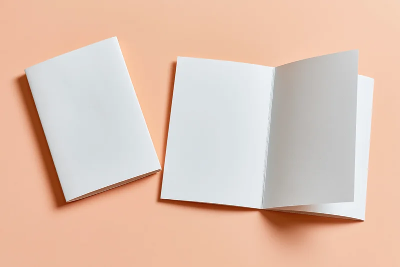 Two blank white bifold brochures one open one closed on peach-orange surface