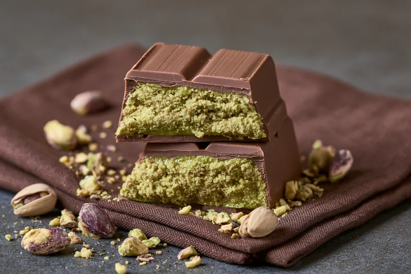 Broken pistachio-filled milk chocolate bar stacked on a brown cloth with scattered nuts and shells