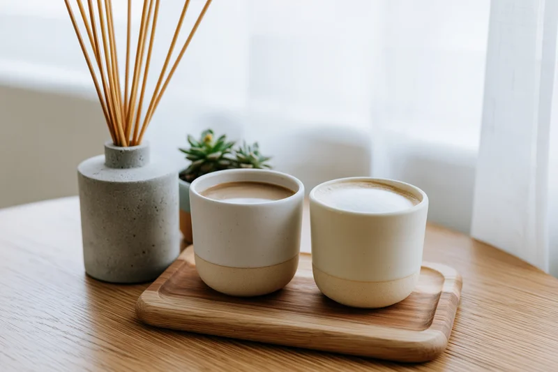 Two ceramic coffee cups on a wooden tray beside a reed diffuser and succulent on a table near a window