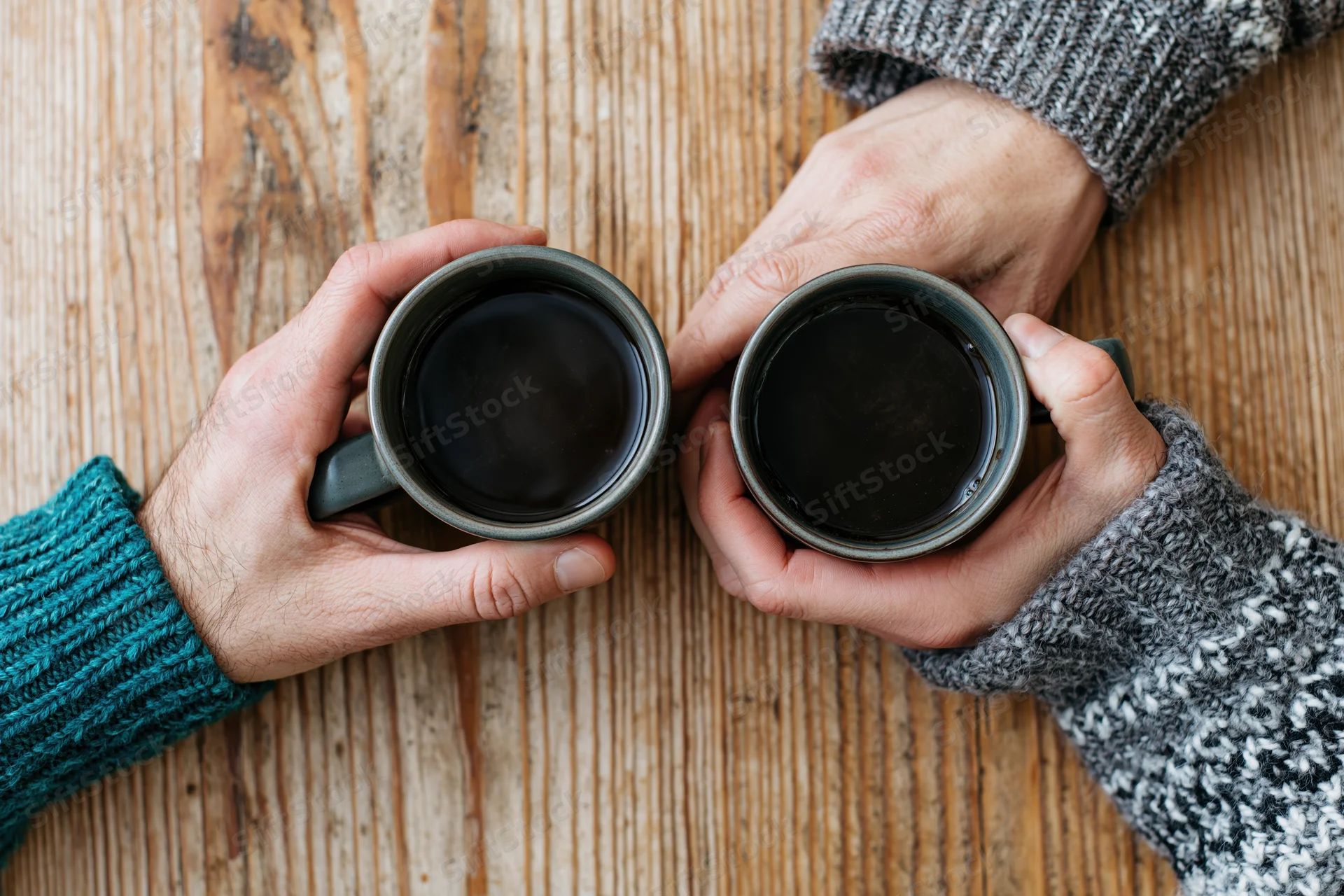 Two hands holding ceramic mugs of black coffee on a wooden table