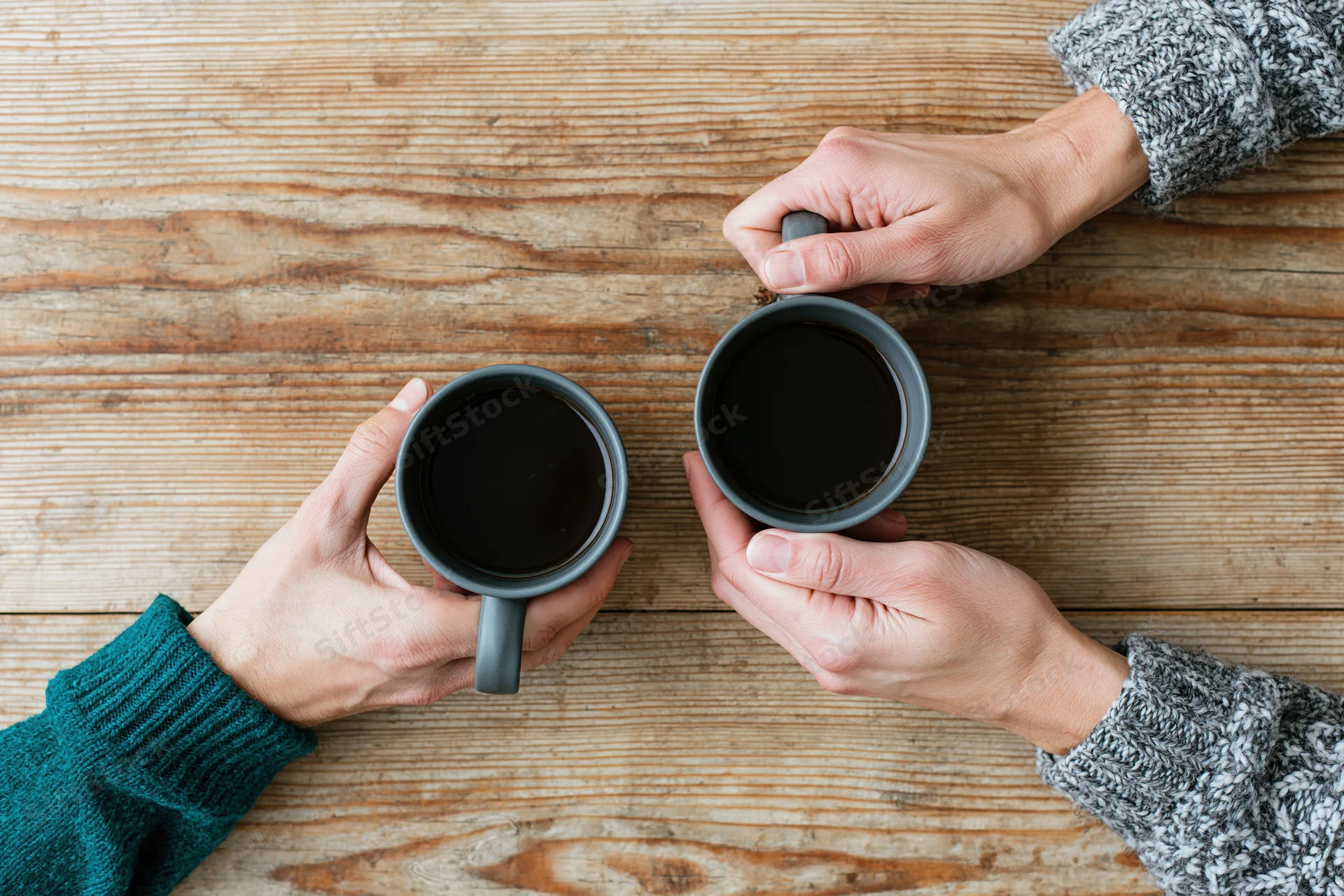 Two pairs of hands holding ceramic mugs of black coffee on a wooden table