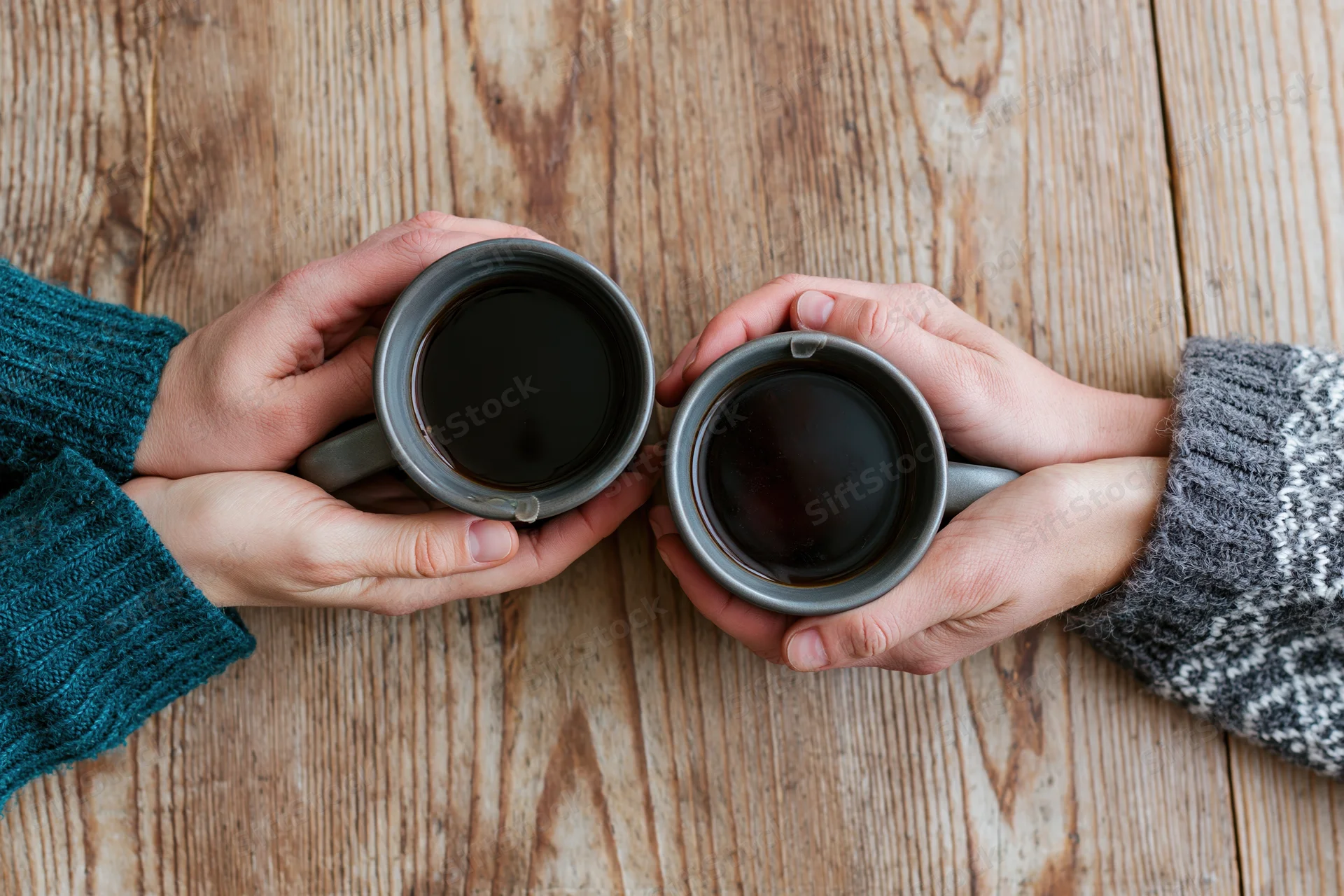 Two pairs of hands holding gray ceramic mugs of black coffee on a wooden table