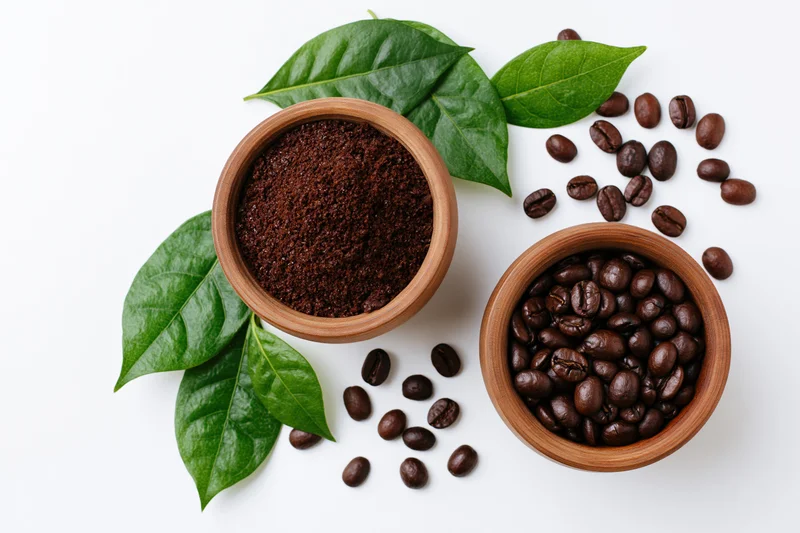 Two wooden bowls with whole and ground coffee beans and green leaves on white background