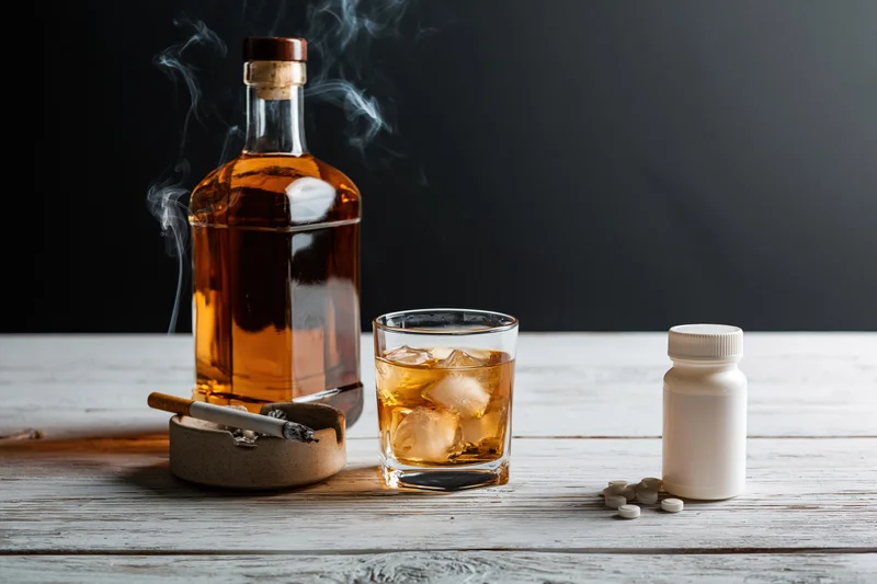 Whiskey bottle and glass with ice beside a smoking cigarette and pills on a white wooden table