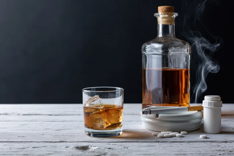 Whiskey glass with ice and bottle on white wooden table beside cigar, ashtray and pills