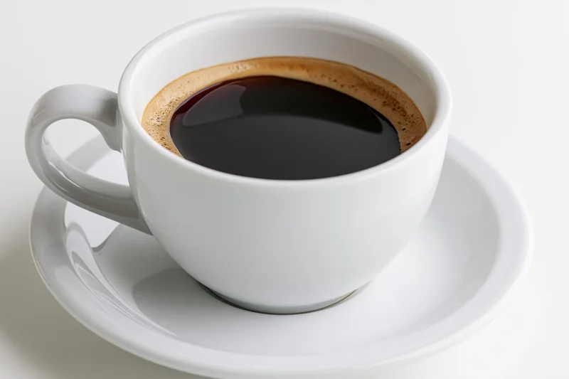 White ceramic coffee cup filled with black coffee and crema on a saucer against a plain white background