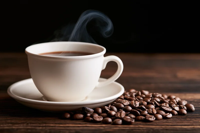 White ceramic coffee cup with steam rising on saucer beside roasted beans on dark wooden table