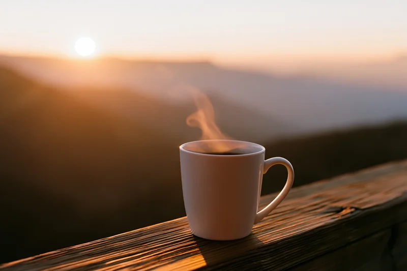 White ceramic coffee mug with steam on wooden railing overlooking mountain sunrise