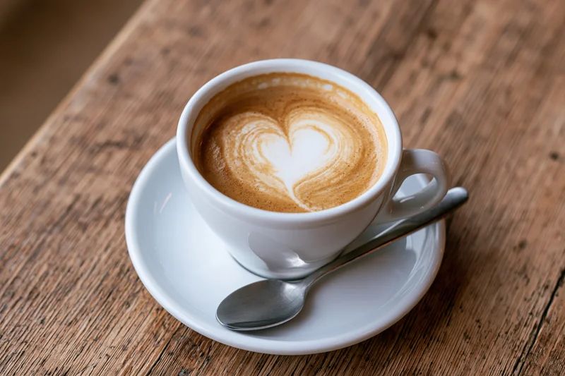 White ceramic cup of latte with heart-shaped foam art on a wooden table with spoon on saucer
