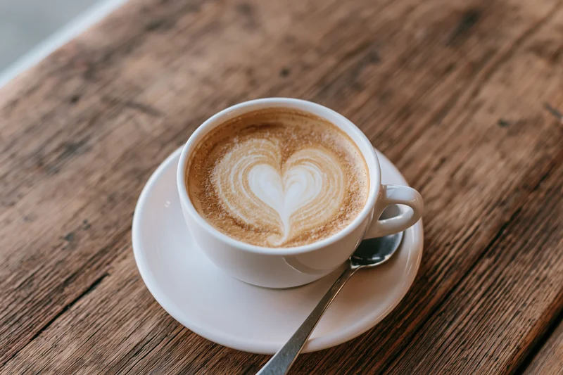 White ceramic cup of latte with heart-shaped milk foam art on wooden table with spoon on saucer