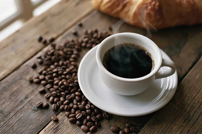 Steaming black coffee in a white ceramic cup on a saucer with scattered coffee beans and a croissant on a wooden table