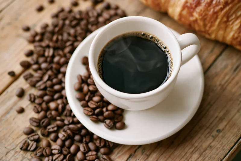 White ceramic cup of steaming black coffee on saucer with coffee beans and croissant on wooden table
