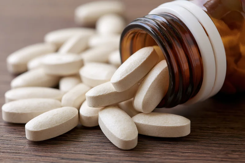 White oval supplement tablets spilling from an amber glass bottle onto a wooden surface