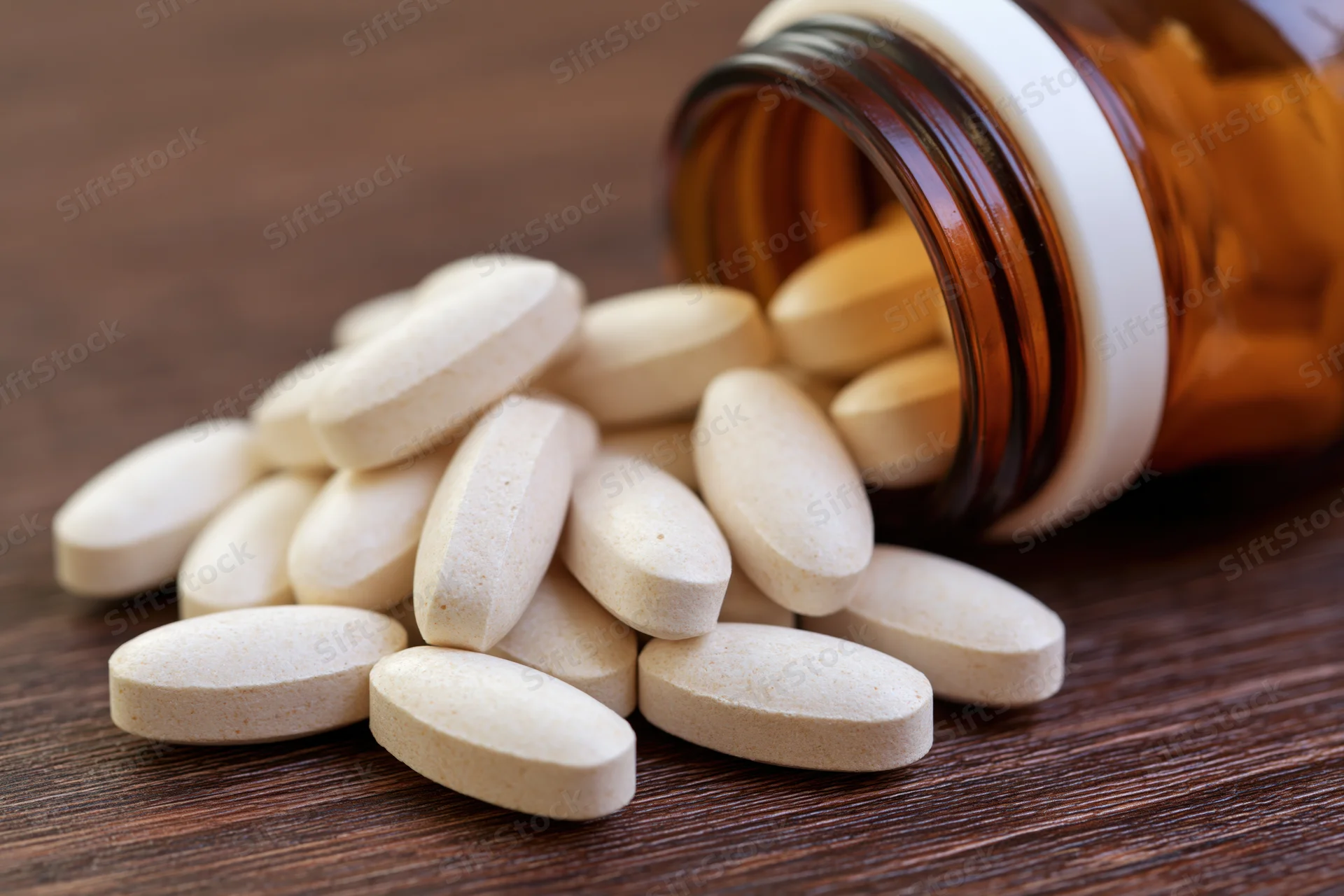 White oval supplement tablets spilling from an amber bottle onto a wooden surface