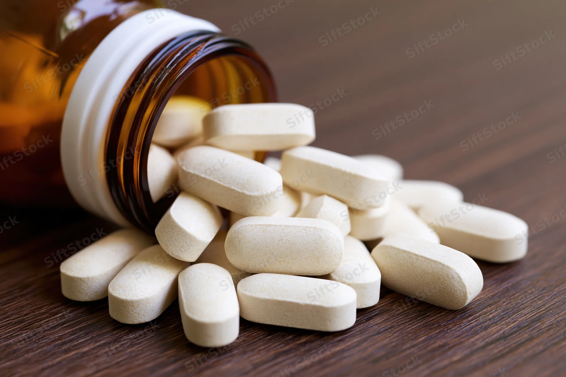 White oval supplement tablets spilling from an open amber glass bottle onto a wooden surface