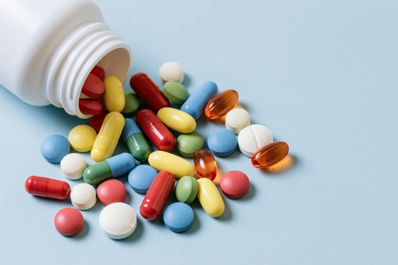 Assorted colorful pills and capsules spilling from a white plastic bottle onto a light blue surface