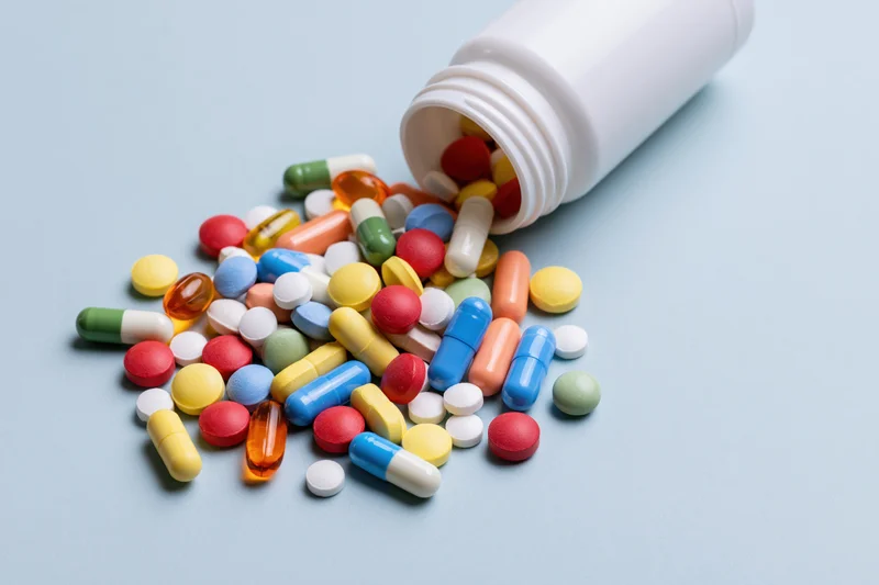 Assorted colorful pills and capsules spilling from a white plastic bottle on a light blue surface