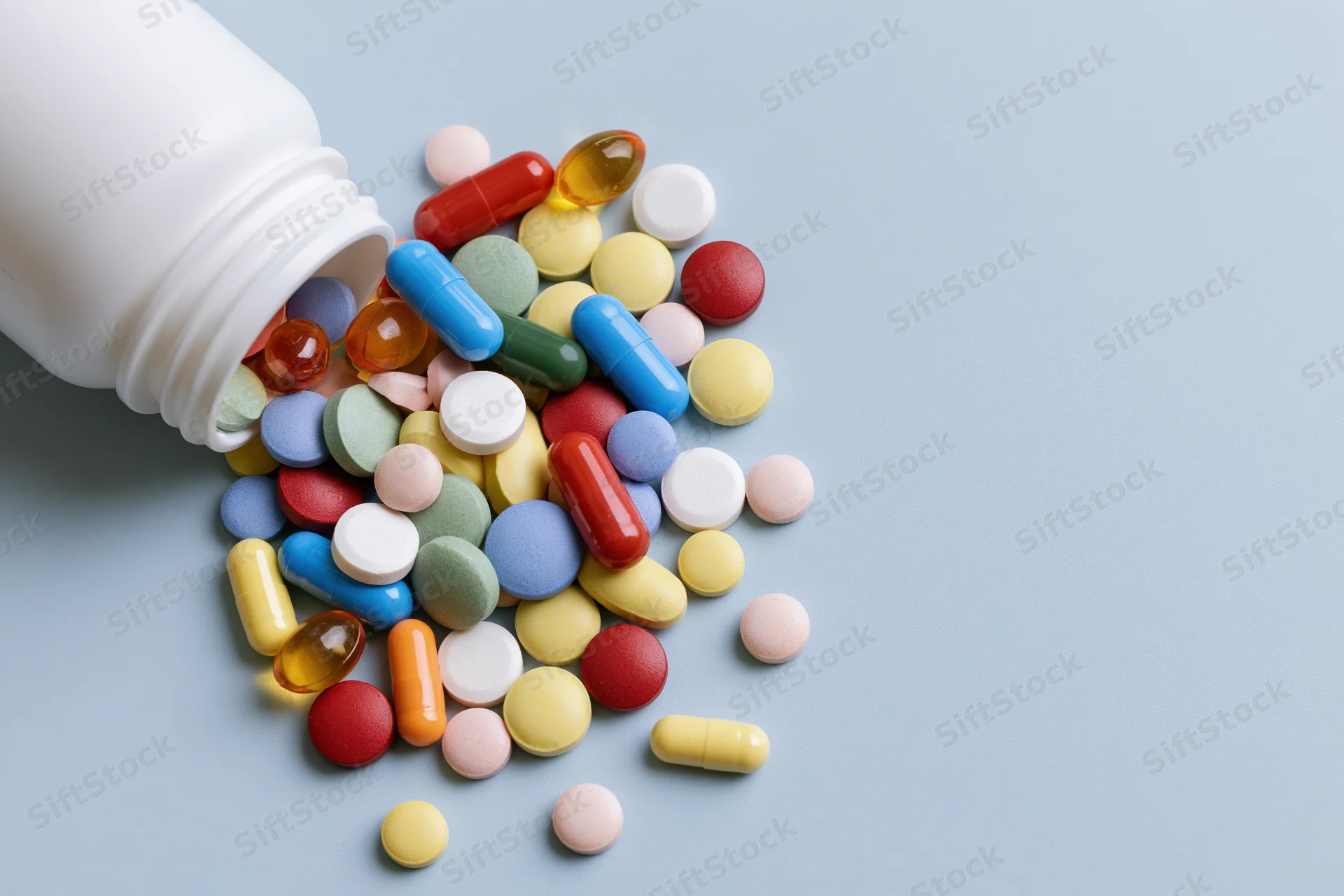 White plastic pill bottle lying on its side with assorted pills spilled onto a light blue surface