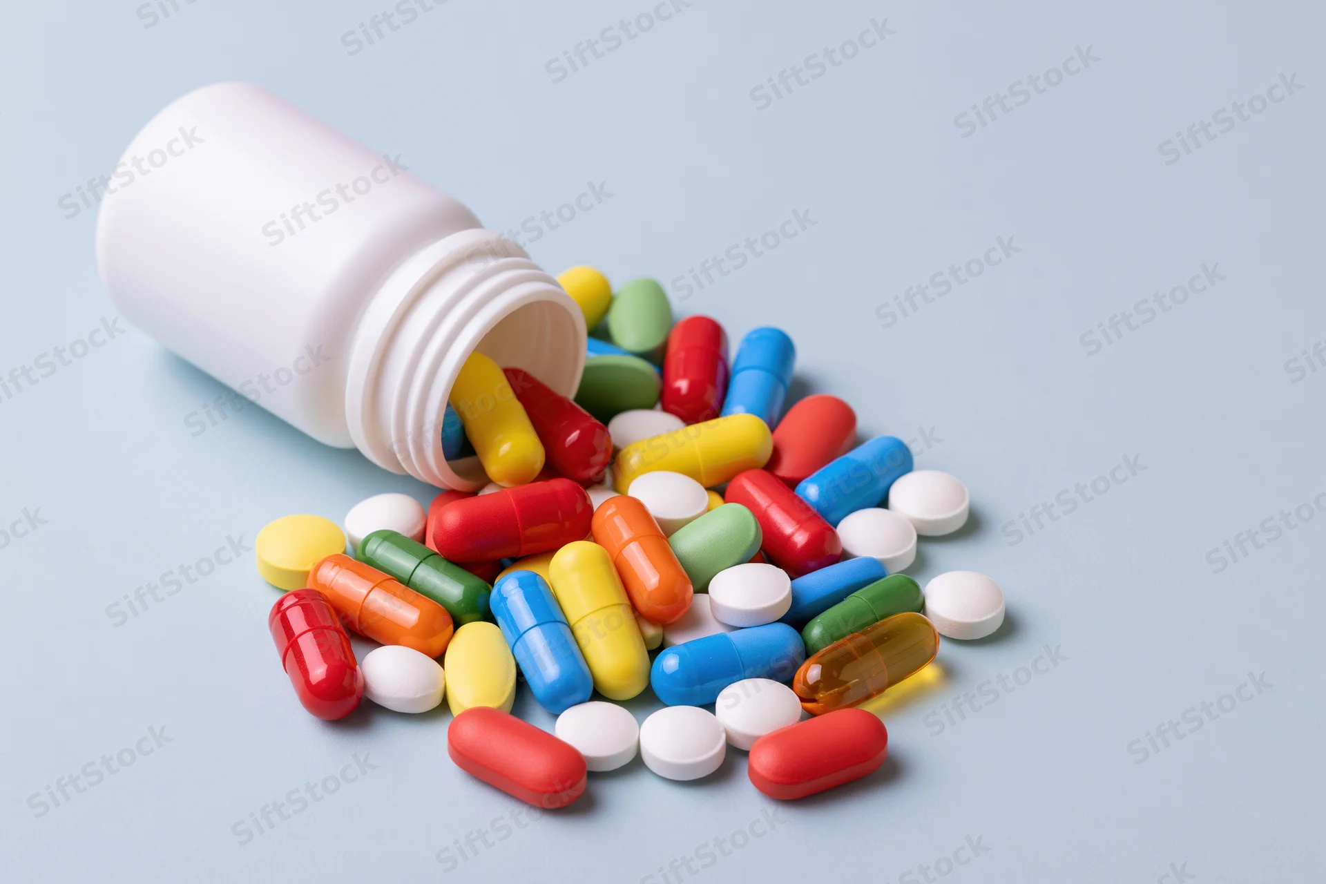 White plastic pill bottle lying on its side with assorted colorful pills spilled onto a light blue surface