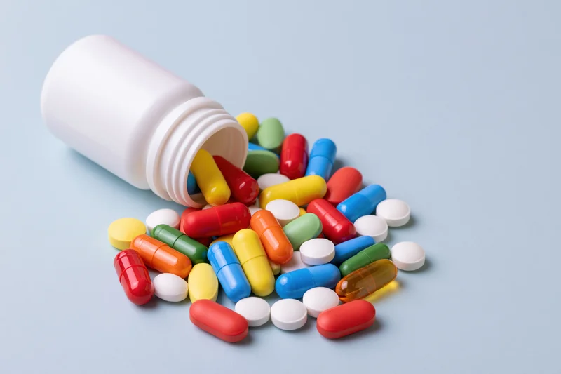 White plastic pill bottle lying on its side with assorted colorful pills spilled onto a light blue surface