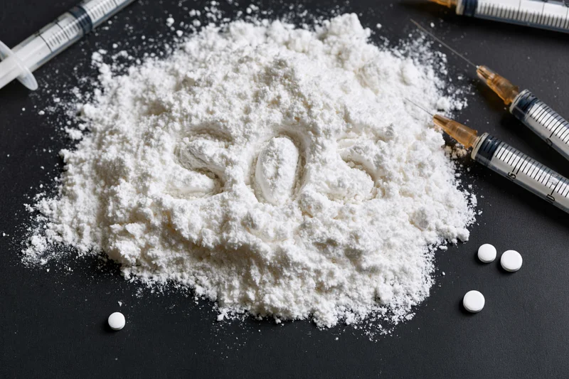 White powder with the letters SOS written in it surrounded by syringes and pills on a black surface
