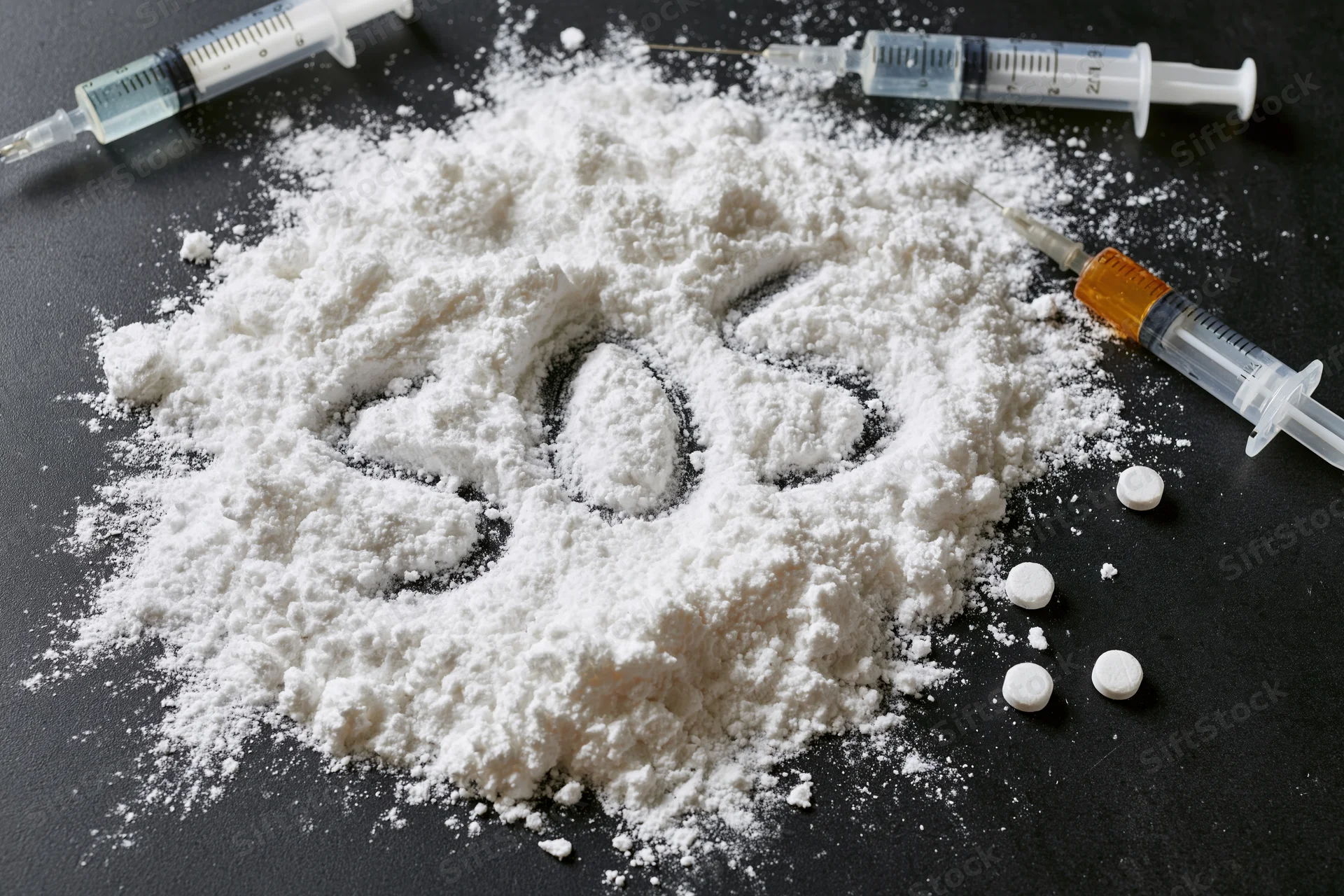 White powder with "SOS" written in it surrounded by syringes and pills on a dark surface