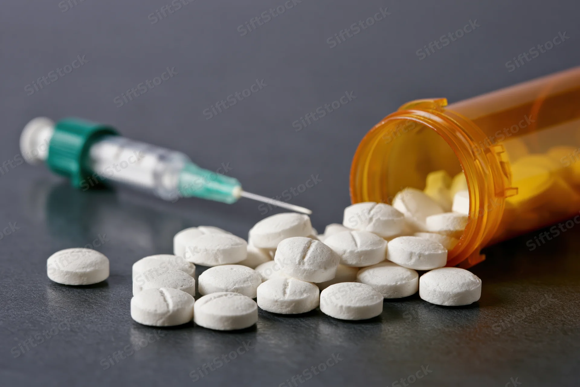 White prescription pills scattered on dark reflective surface with syringe and orange bottle nearby