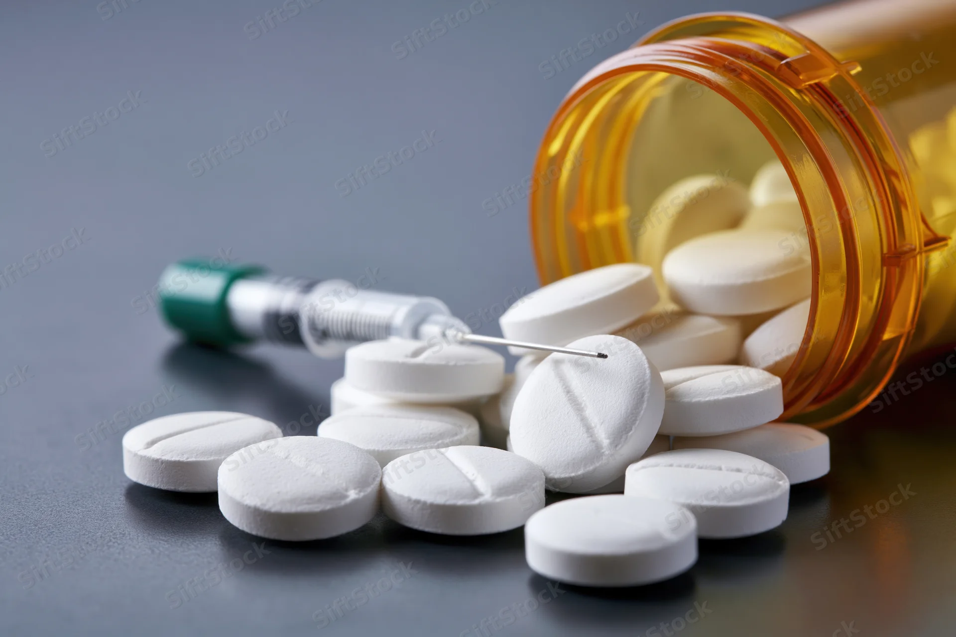 White prescription pills scattered on dark reflective surface with syringe and amber bottle nearby