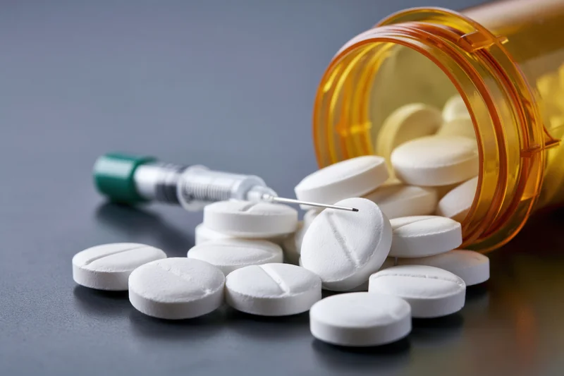 White prescription pills scattered on dark reflective surface with syringe and amber bottle nearby