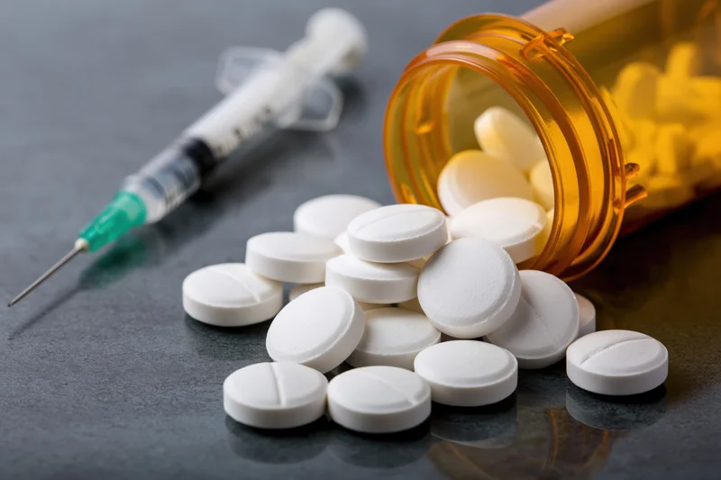 White prescription pills scattered on dark reflective surface with syringe and amber bottle nearby