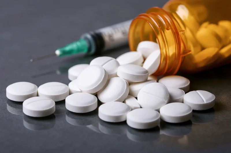 White prescription pills scattered on dark reflective surface with syringe and orange bottle nearby