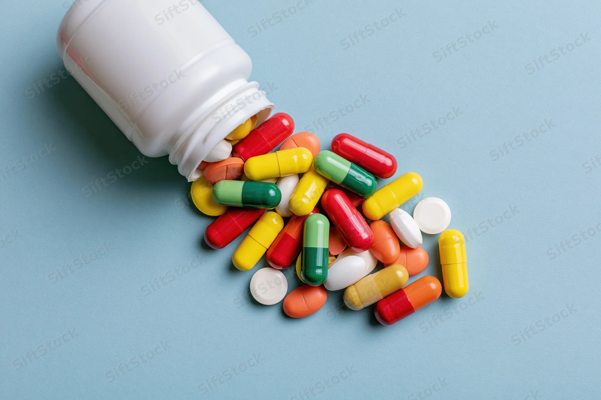 Colorful pills and capsules spilled from a white bottle on a blue surface