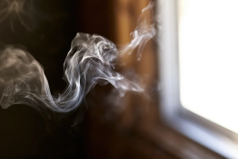 Wispy white smoke curling and dispersing in mid-air near a window indoors