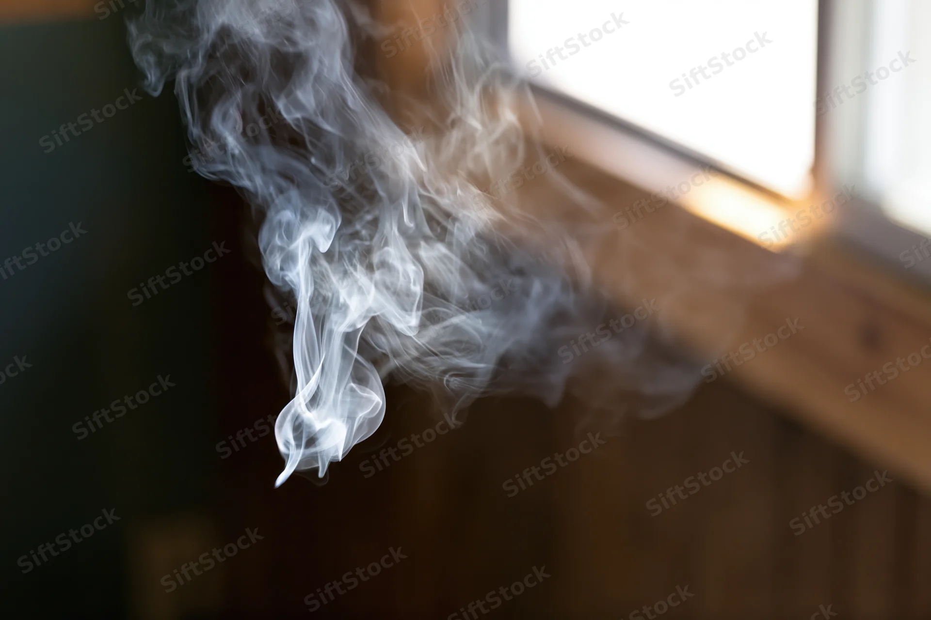 Wispy white smoke curling and dispersing near a window sill in a dimly lit room