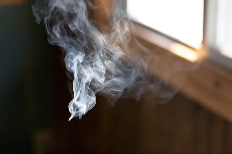Wispy white smoke curling and dispersing near a window sill in a dimly lit room