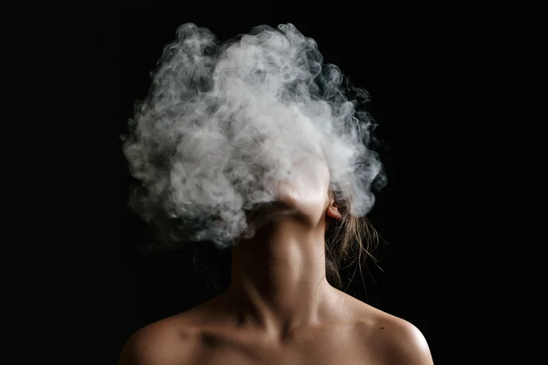 Woman exhaling dense white smoke against black background, bare shoulders visible, head tilted up