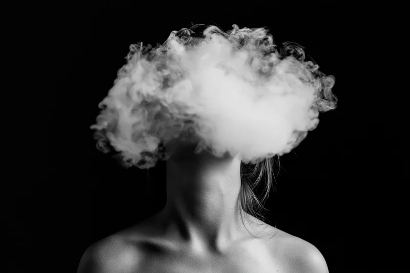 Woman exhaling dense smoke cloud obscuring her head against black background