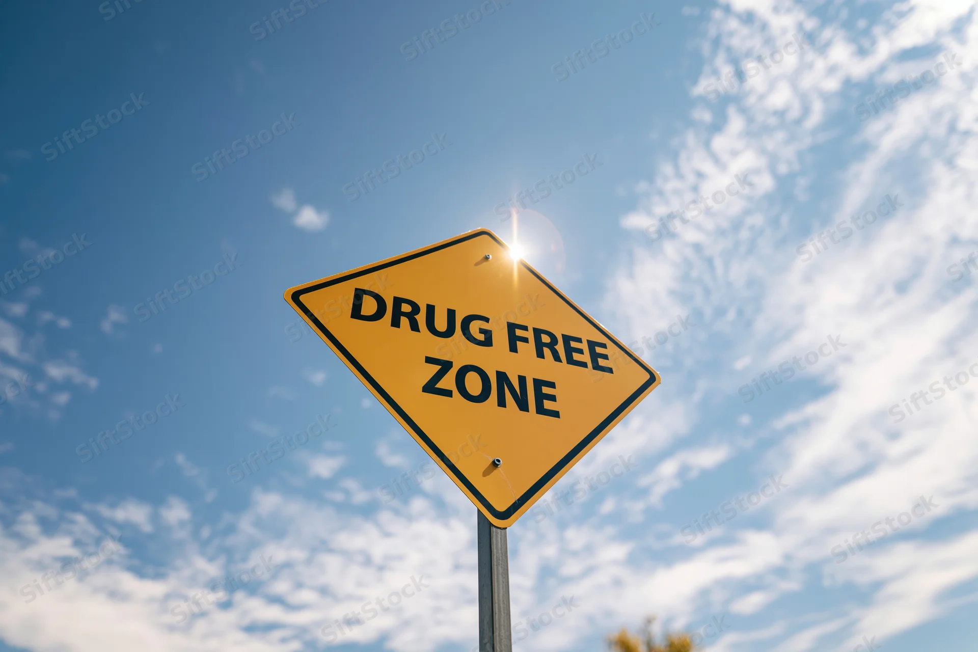 Yellow diamond-shaped drug free zone sign on a metal post against a blue sky with scattered clouds and sunlight