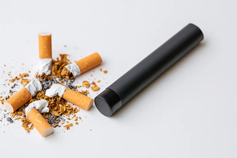 Black vape pen lying beside crushed cigarettes and tobacco debris on white surface