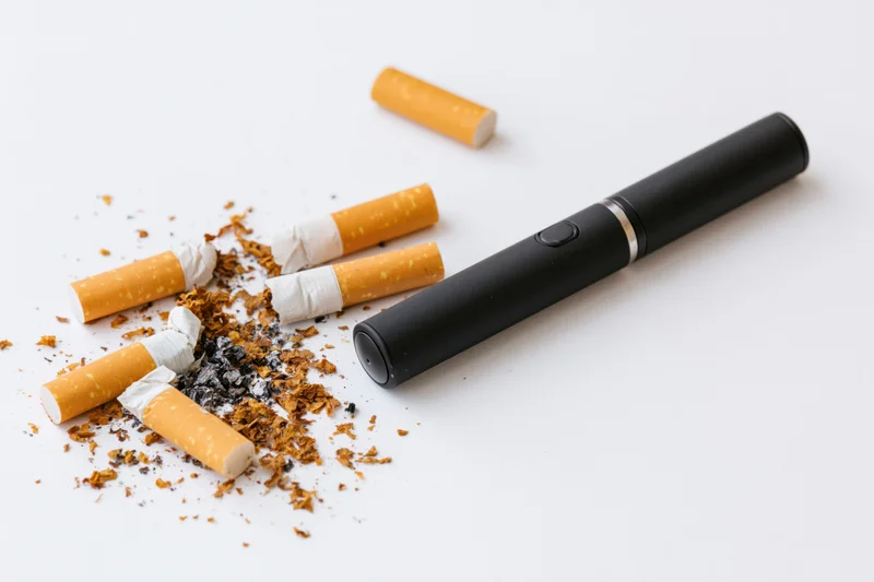 Black e-cigarette vape pen lying beside crushed cigarettes and tobacco on white surface