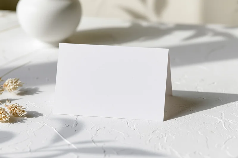 Blank white folded tent card standing on textured surface with dried grass and soft shadows