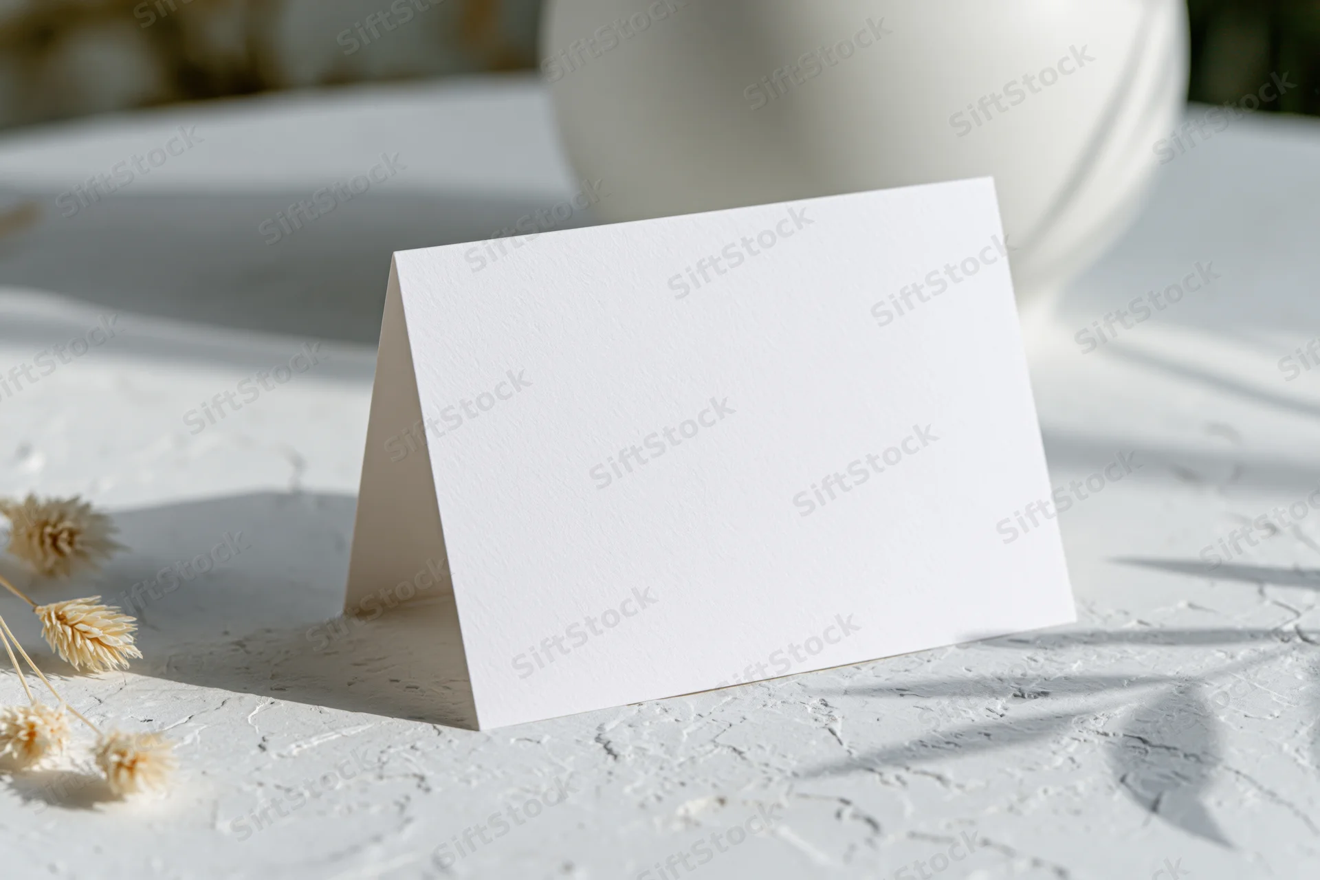 Blank white folded tent card standing on textured white surface with dried flowers and soft shadows