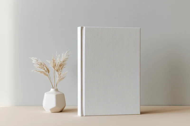 Blank white hardcover book standing upright next to a small vase with dried pampas grass on a beige surface against a light gray wall