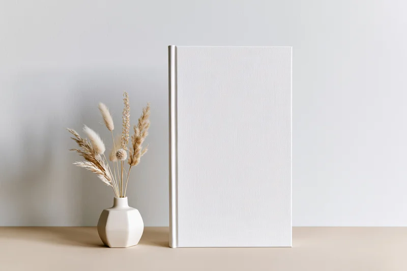 Blank white hardcover book standing upright on beige surface beside vase with dried pampas grass