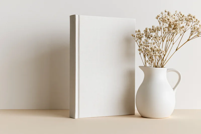 Blank white hardcover book standing upright next to a white ceramic vase with dried gypsophila flowers on a beige surface
