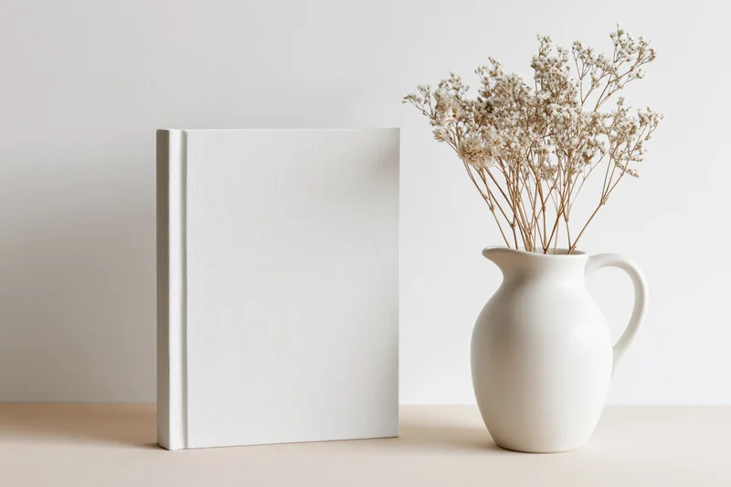 Blank white hardcover book standing next to a white ceramic pitcher with dried baby's breath flowers on a beige surface