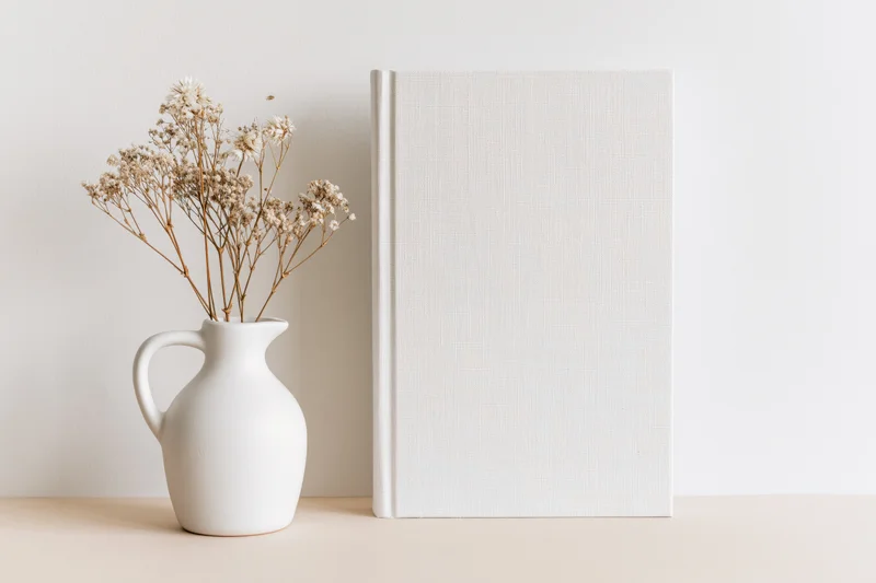 Blank white hardcover book standing upright next to a white ceramic vase with dried flowers on a beige surface