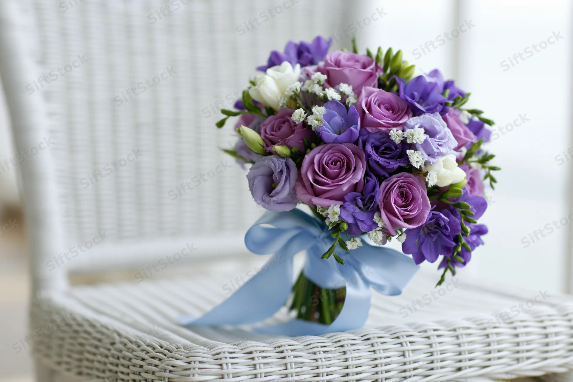 Bridal bouquet of purple roses and lavender lisianthus with blue ribbon on white wicker chair