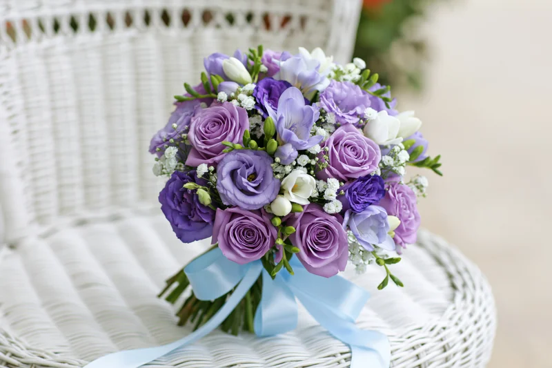 Bridal bouquet of purple roses, lavender lisianthus and freesia resting on a white wicker chair